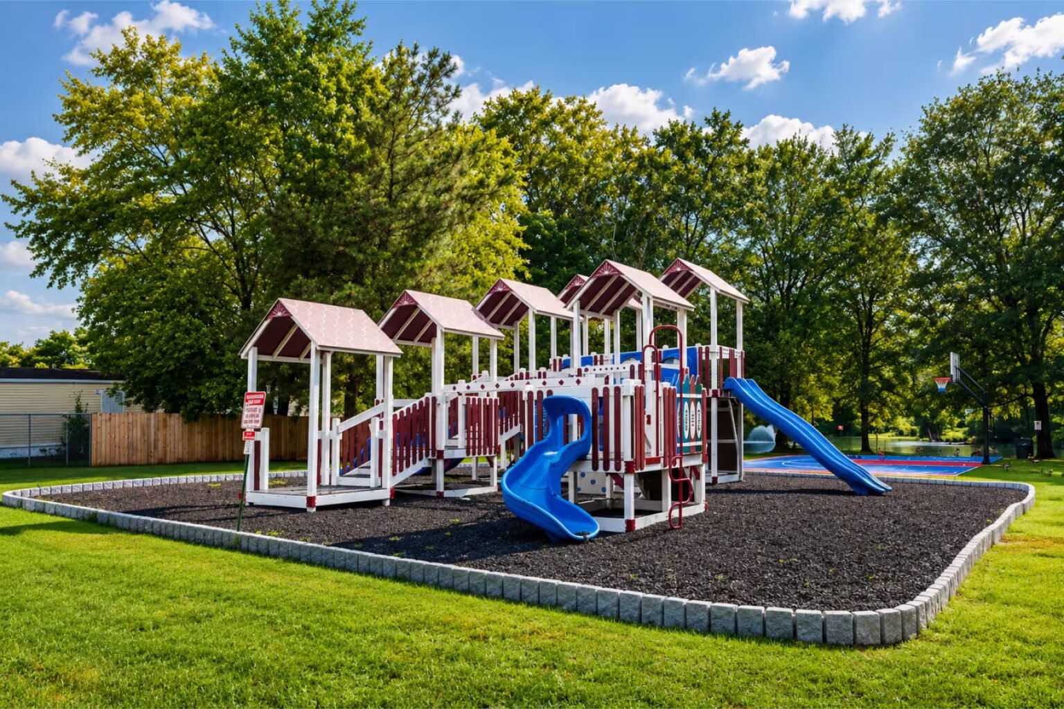 Children's playground featuring slides, climbing structures, and swing set on wood chips, surrounded by green lawn and trees.