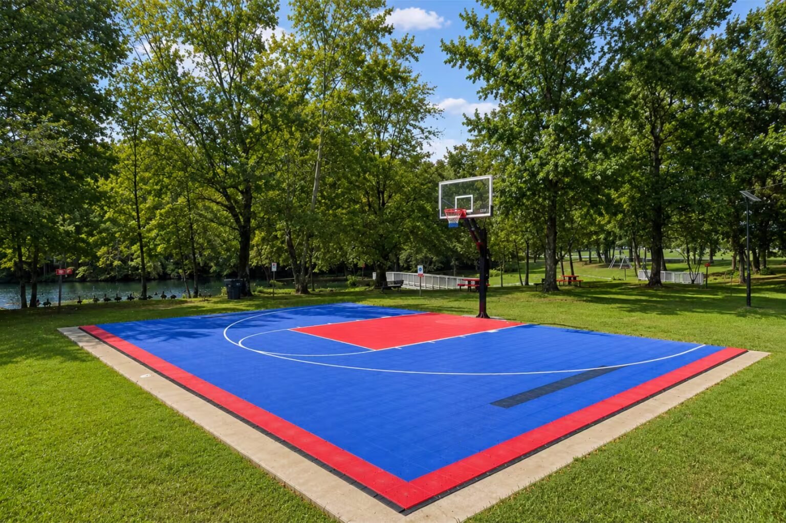 Basketball hoop on a colorful blue-and-red outdoor court surrounded by grass and green trees.