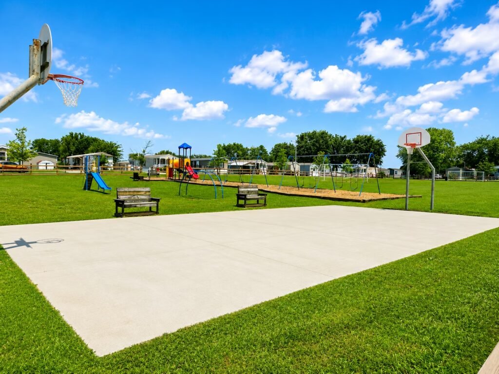 Empty park lot bordered by grass, with colorful playground equipment under bright cloudy sky.