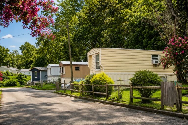 Static caravans and trailers parked along a paved lane surrounded by green trees on a sunny day.