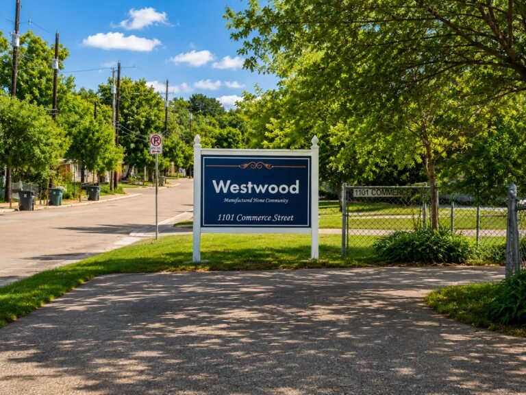 Blue welcome sign reading "Welcome" at roadside entrance framed by lush green trees under sunny sky.