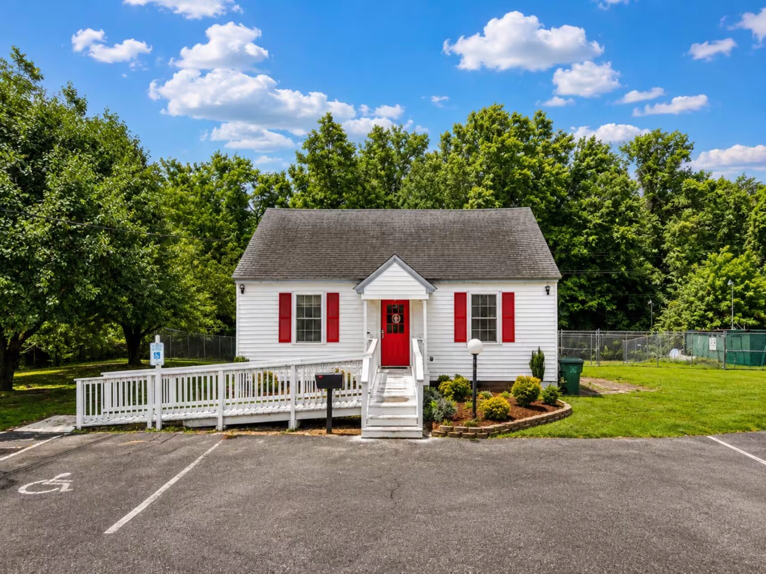 White cottage-style building with a bright red front door and shutters beneath a partly cloudy blue sky.