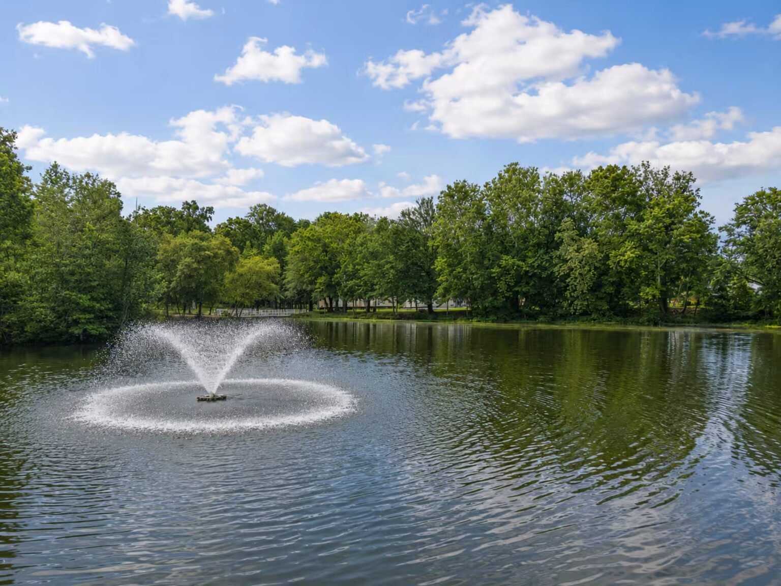 A fountain sprays water in a tranquil pond surrounded by lush green trees beneath a partly cloudy sky.