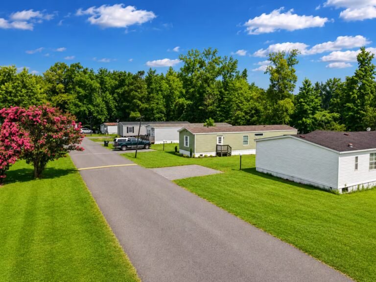 Single-wide manufactured homes line both sides of a paved road beneath a partly cloudy blue sky.