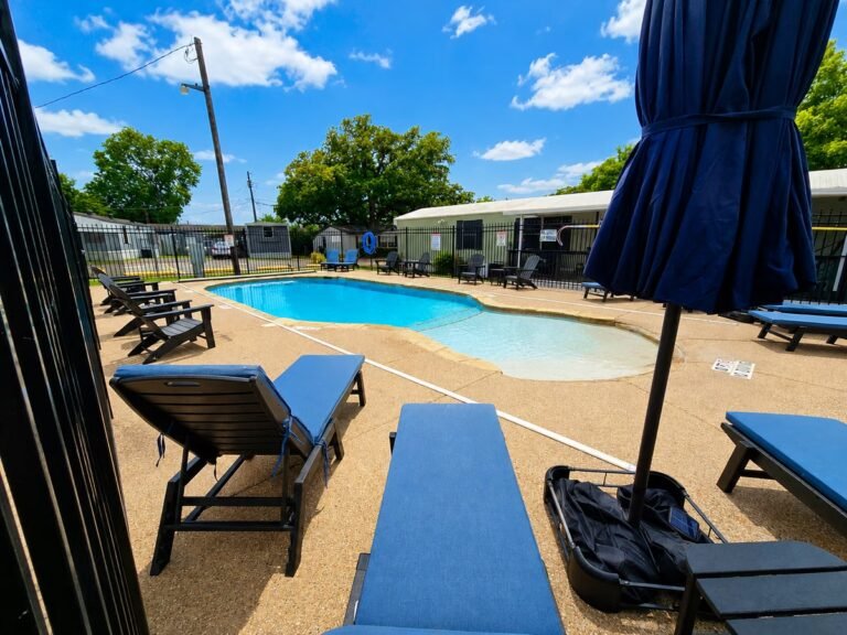 Resort swimming pool surrounded by lounge chairs beneath blue shade umbrellas on a sunny day.