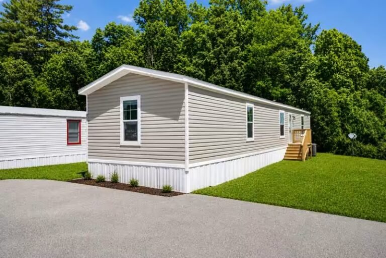 White single-wide manufactured home with grey metal roof, set on a green lawn with dense trees behind it.