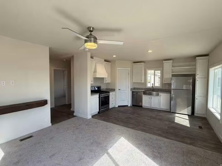 Empty beige-carpeted living area opening to a kitchen with white cabinets, wood accents, and glass slider.