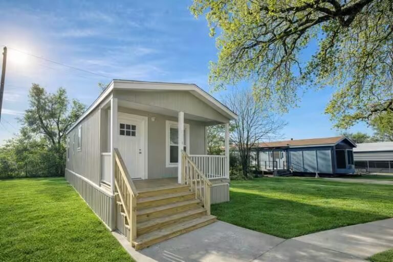 White single-wide manufactured home with covered front porch and steps, set on a green lawn beneath blue skies.