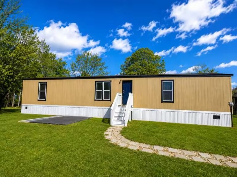 Tan single-wide mobile home with blue front door on grassy lot beneath bright partly cloudy sky.
