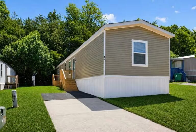 Tan single-wide manufactured home with white skirting, ramp entry, and walkway on a green lawn.