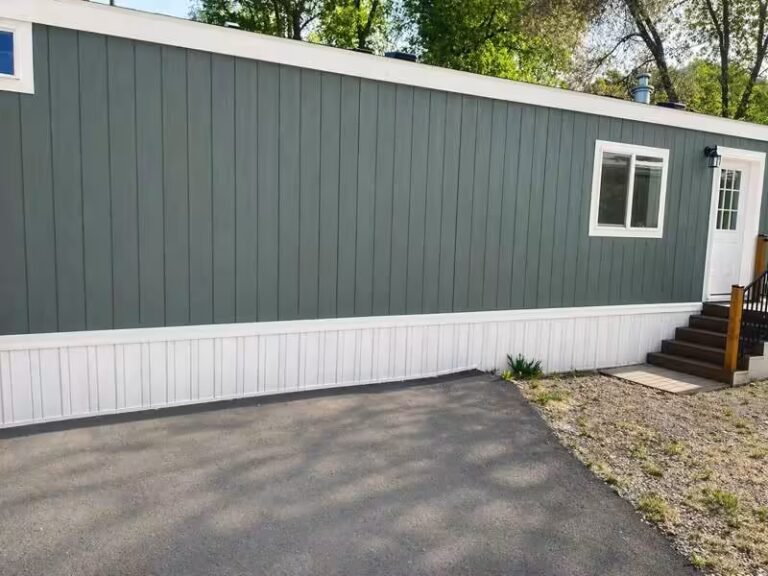 A green-sided mobile home with white front door and small round table near its entrance.