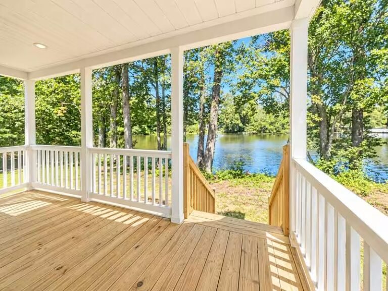 Covered wooden porch with white railings overlooking a calm lake surrounded by lush green trees.