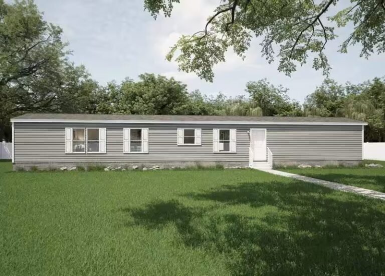 White single-wide manufactured home with dark window shutters sits on a grassy lot beneath tall leafy trees.