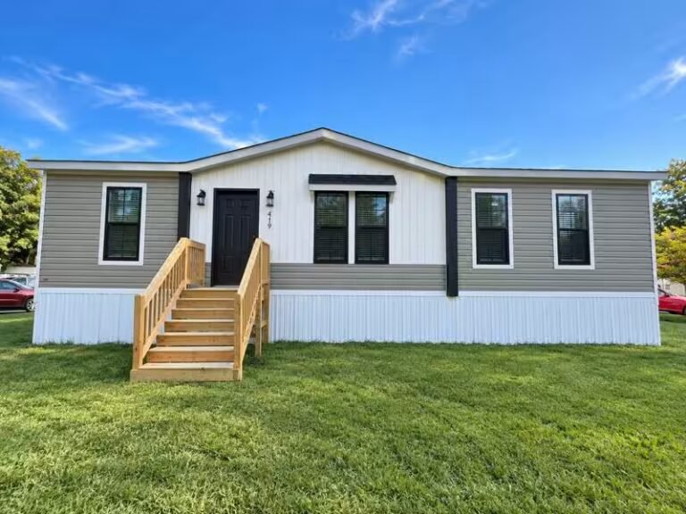Gray and white single-wide mobile home with black front door, dark shutters, and wooden entry ramp.