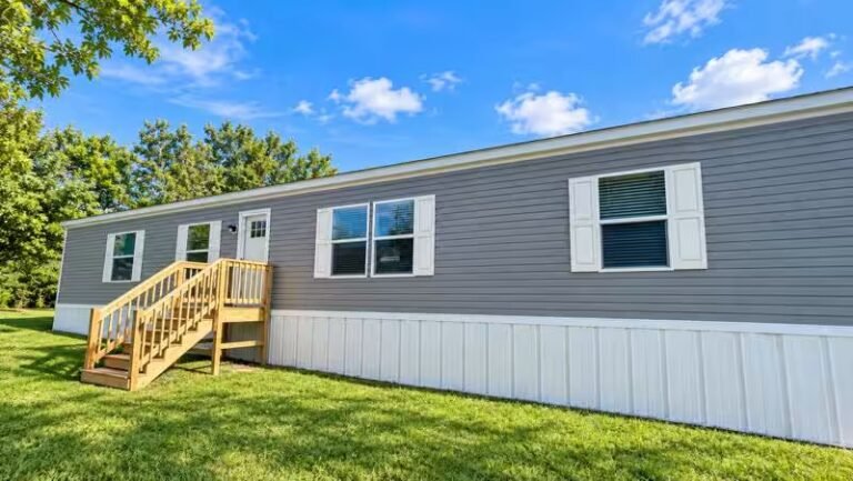 Single-wide gray manufactured home with white skirting and a wooden accessibility ramp and stairs.