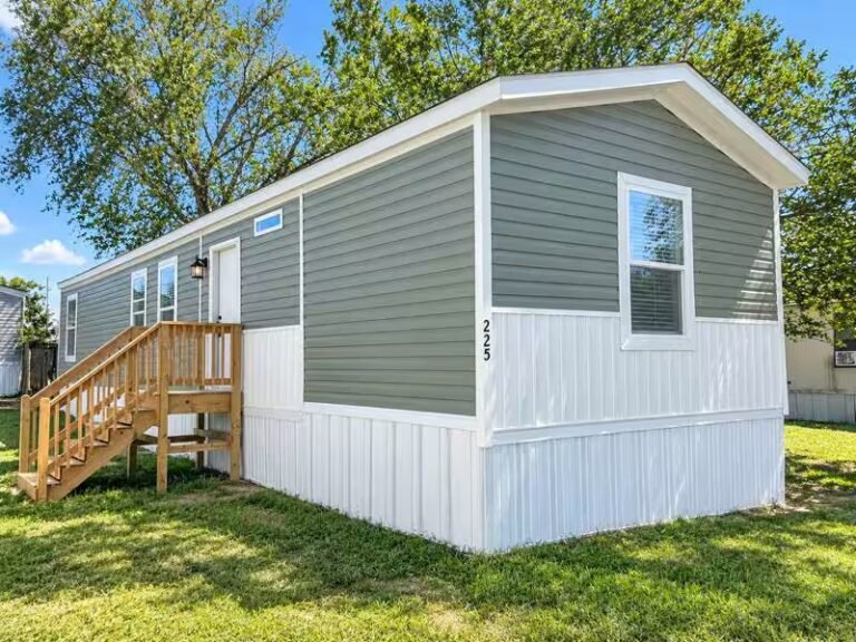 Single-wide manufactured home in sage and white with wheelchair ramp on front porch under blue sky.