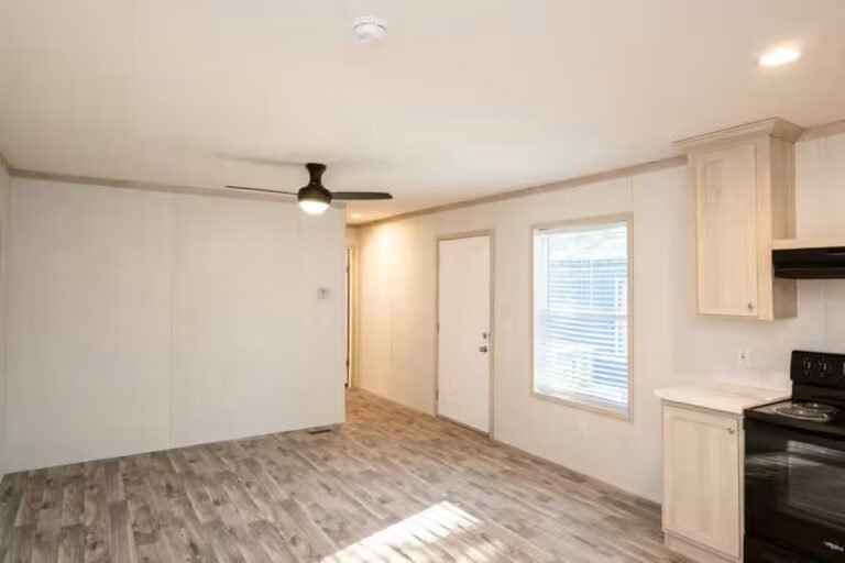 Empty room with wood floors, ceiling fan, window, and kitchen area with cabinets and black stove