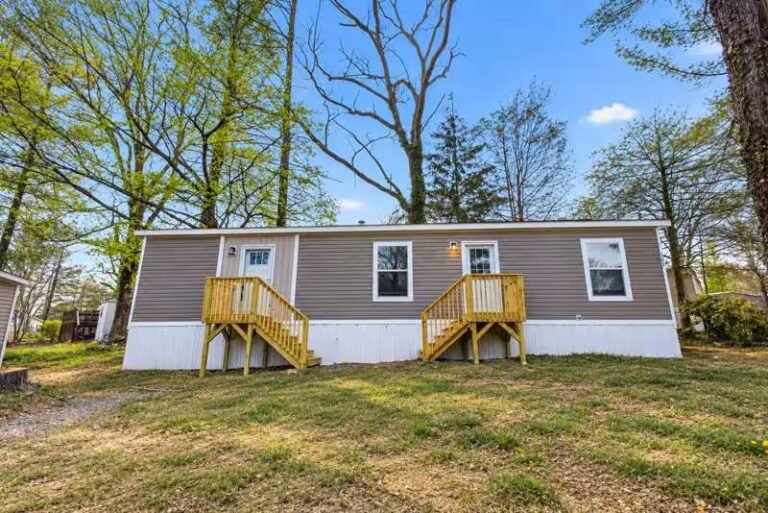Tan single-wide manufactured home with yellow porch steps on a green lawn surrounded by bare trees under blue sky.