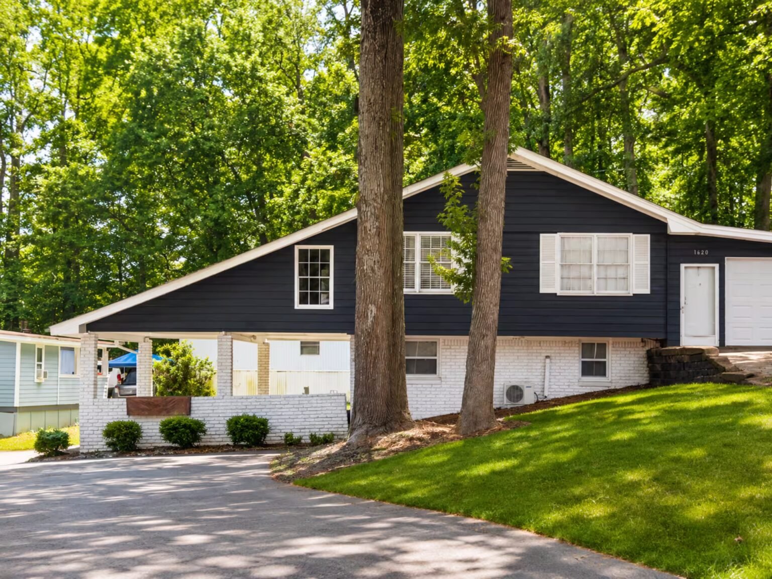 A midcentury-style home with a black paneled facade above its white first floor is nestled among tall leafy green trees