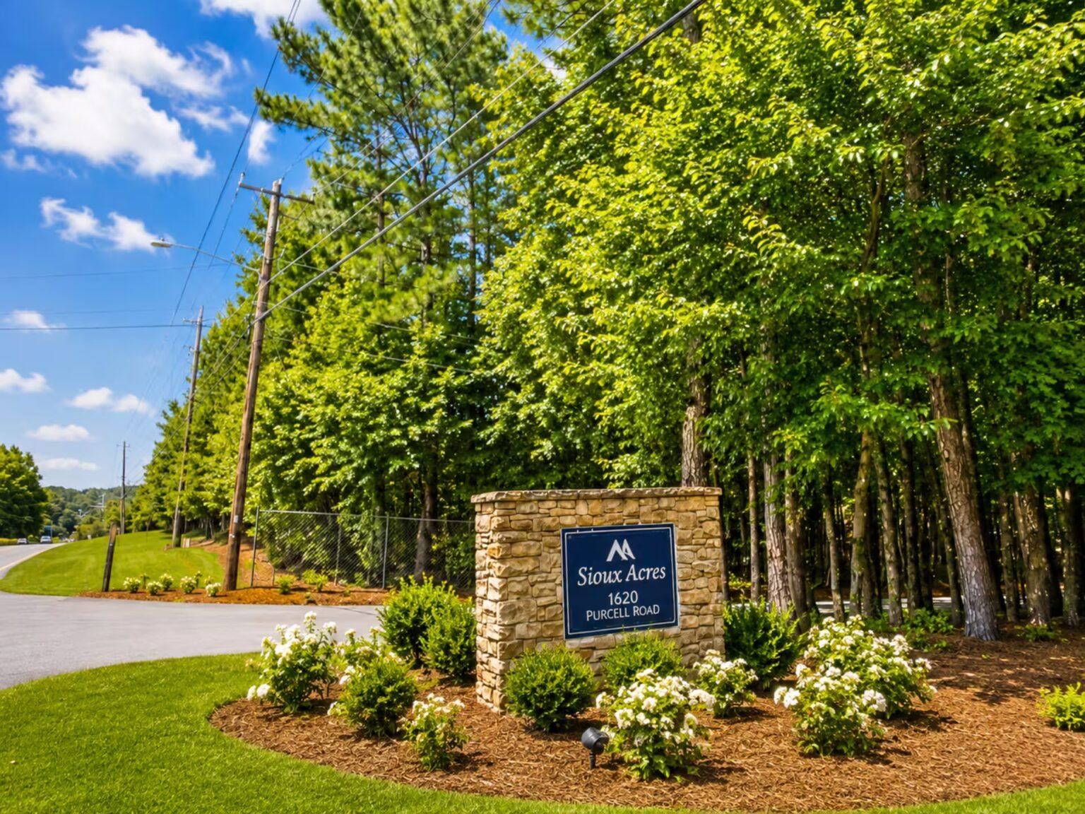 Blue community entrance sign nestled among green bushes and tall pine trees along a curving roadside landscape.