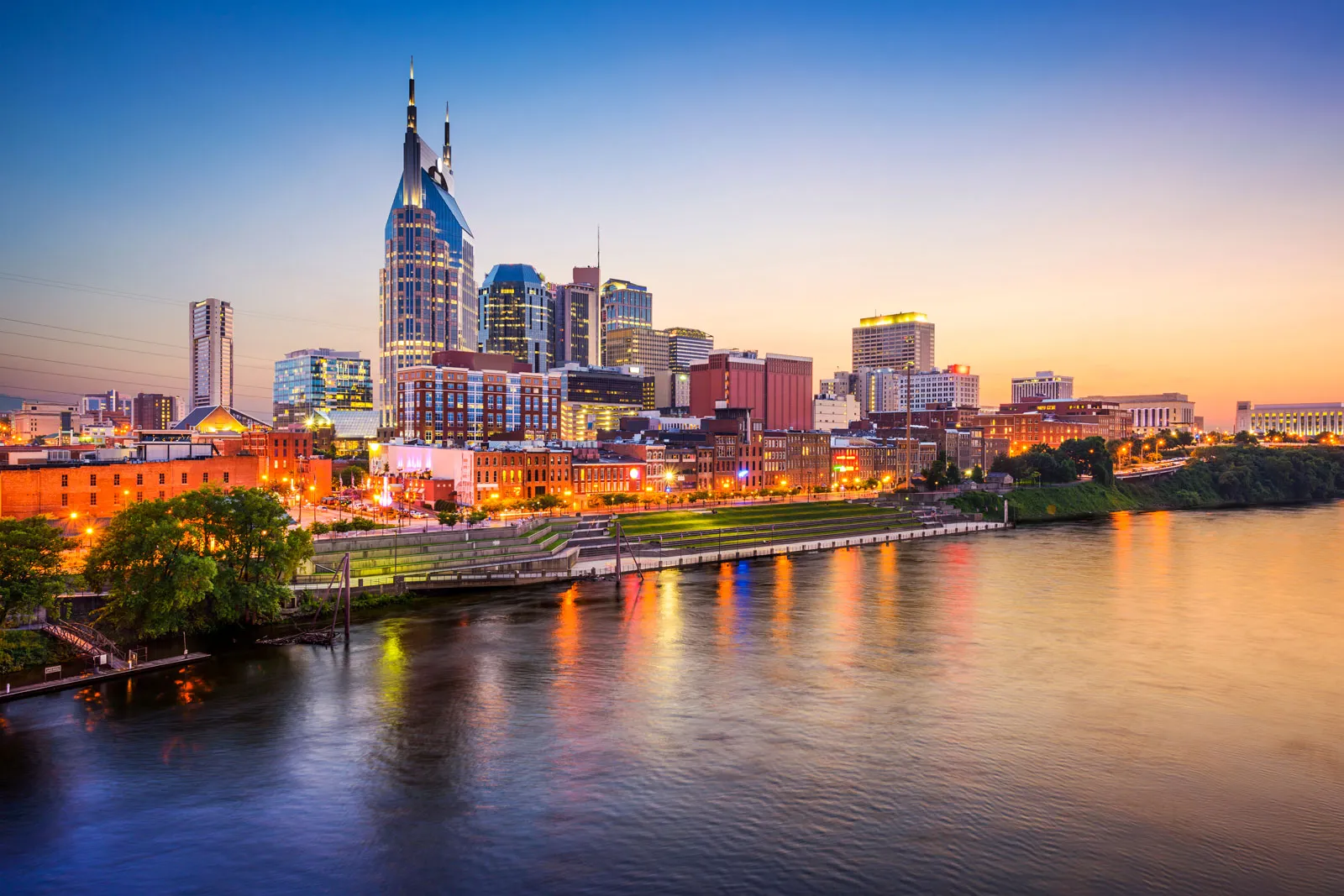 City skyline glowing at sunset beside a calm river reflecting warm lights from downtown buildings.