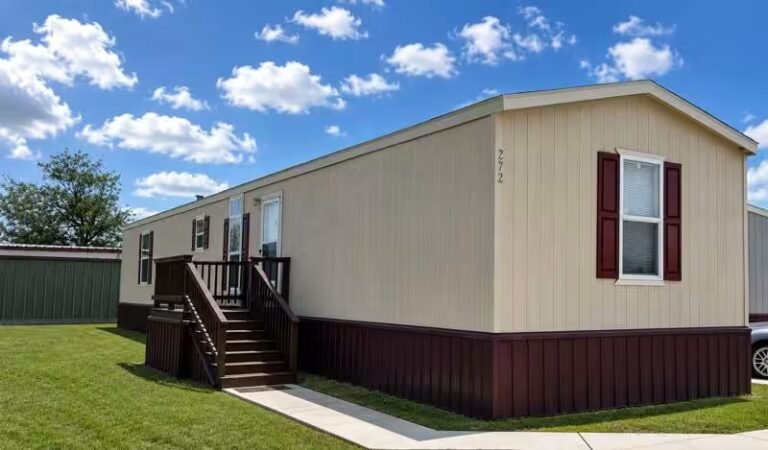 White single-wide mobile home with red-trimmed windows and entry stairs under a bright, cloud-filled sky.