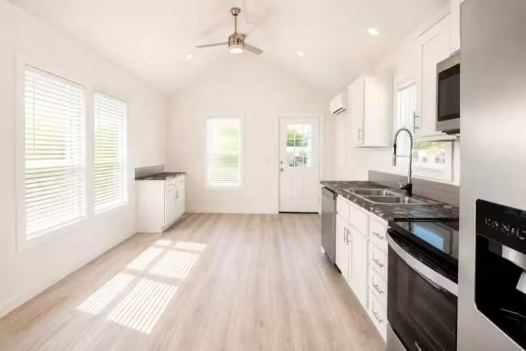 Bright kitchen and living space with white cabinetry, black stove, ceiling fan, large windows, light wood flooring.