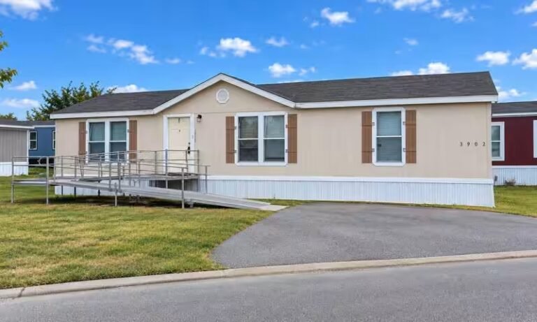 Beige manufactured home with white shutters and windows beside a paved asphalt driveway under partly cloudy skies