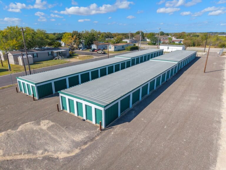 Rows of teal self-storage rental units with gray roofs beside a gravel driveway near homes.
