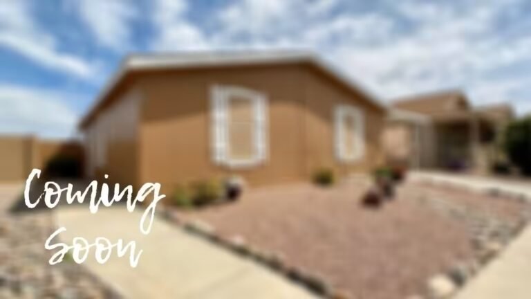 Tan manufactured home on gravel lot under blue sky; "Coming Soon" sign displayed in foreground.