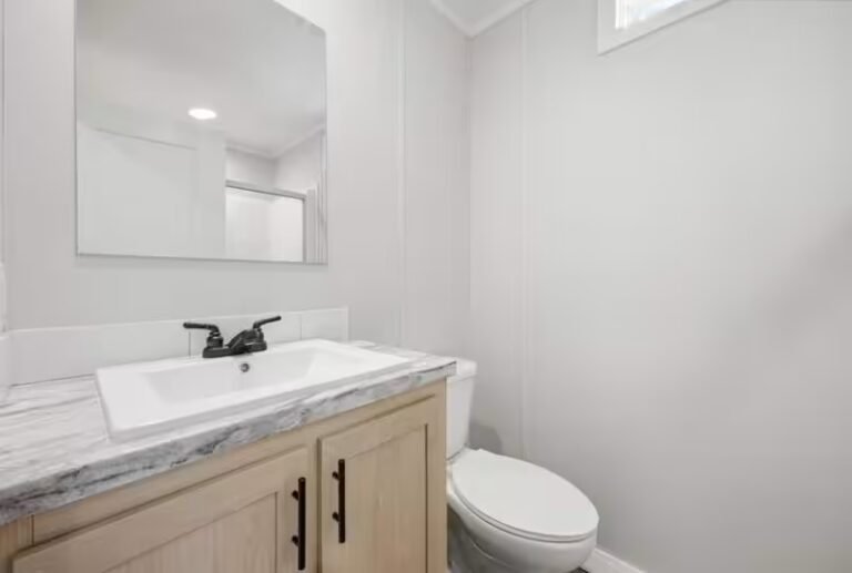 Bathroom corner with white wall, silver-framed mirror over light wood vanity beside a white toilet.