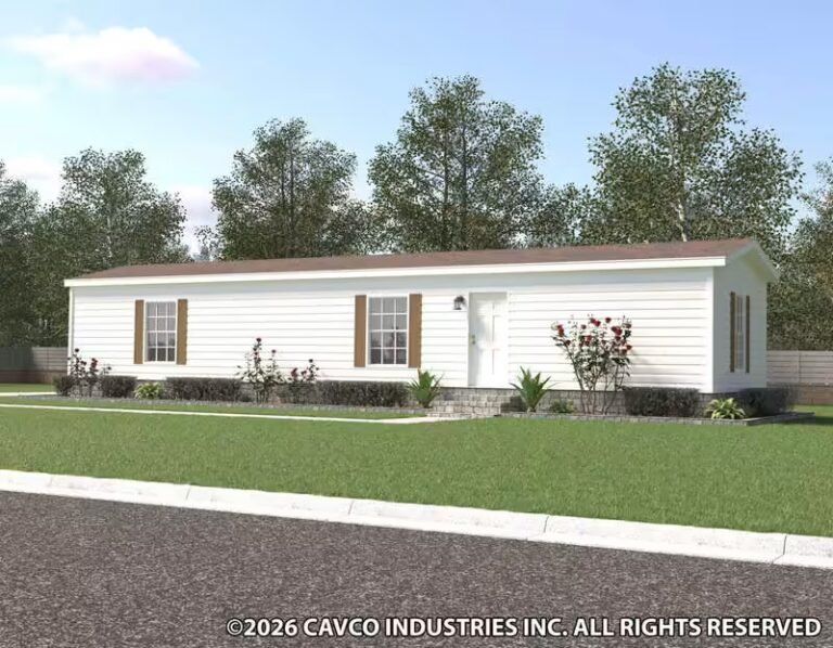 White single-wide mobile home with front porch on green lawn, bordered by mature trees under clear sky.