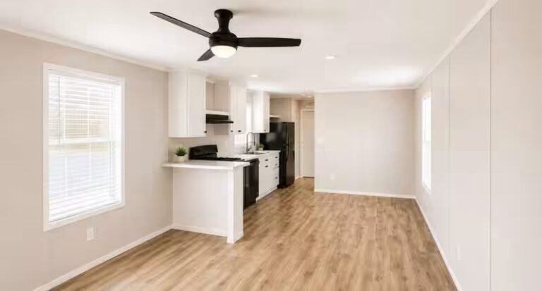 Bright vacant room with wood-look flooring and a ceiling fan opening into a small white kitchen.