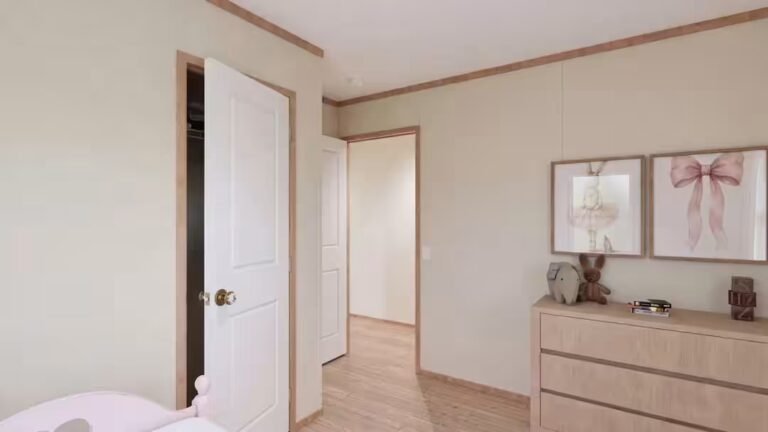 Empty bedroom corner with wooden dresser topped by framed print beside an open doorway leading to a hall.